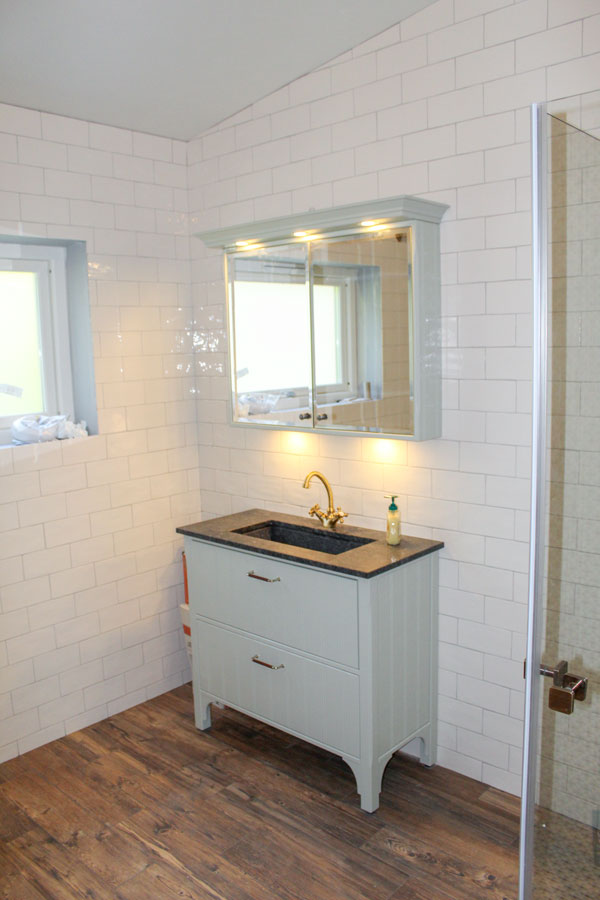 Kakel i Värmland - badrumskomod i vitt, badrumsspegel, handfat, vita kakelplattor på väggen, mörkt trägolv